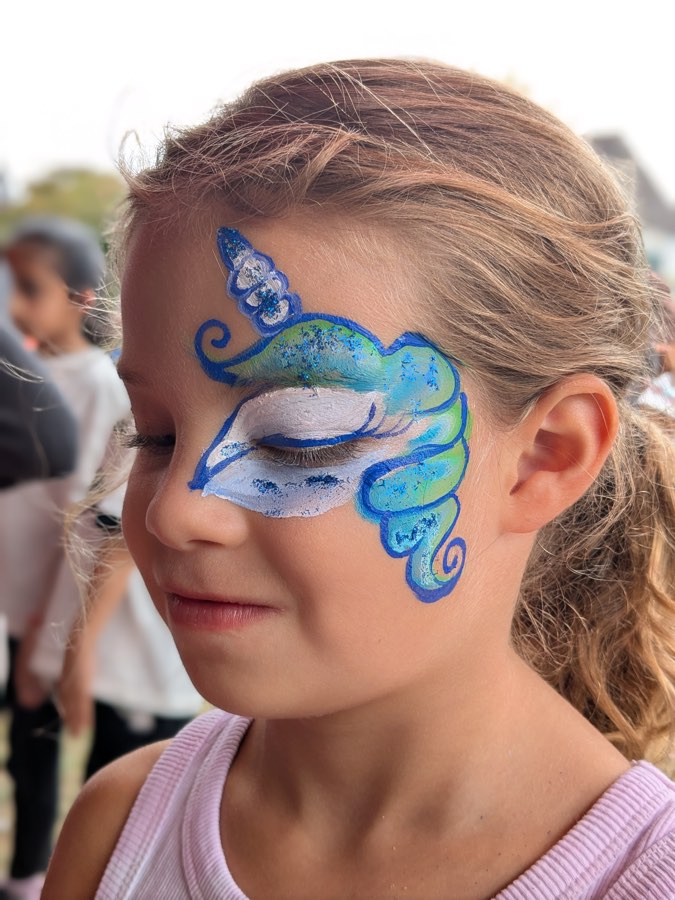 Child with a blue and turquoise unicorn face painting with a gold horn and swirls around one eye