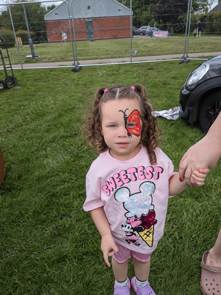 A toddler with a red butterfly cheek painting at an outdoor family event